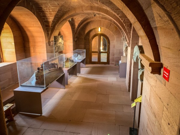 Der Felsblock, der den Palas trägt, ist in der Höhe geteilt: Die hier betrachtete östliche Hälfte des Palasunterschosses liegt auf dem Niveau des ersten Turmgeschosses. Der Raum dient museal der Präsentation mehrer Burgmodelle und Bogensteine . Dauerausstellung Burgmodelle im Palasuntergeschoss des Trifels